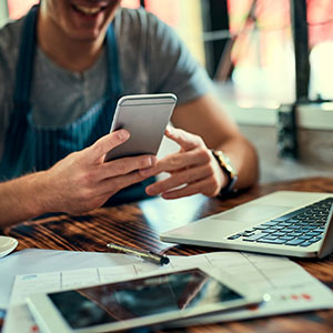 Business man in apron using phone sitting at table with laptop and papers.